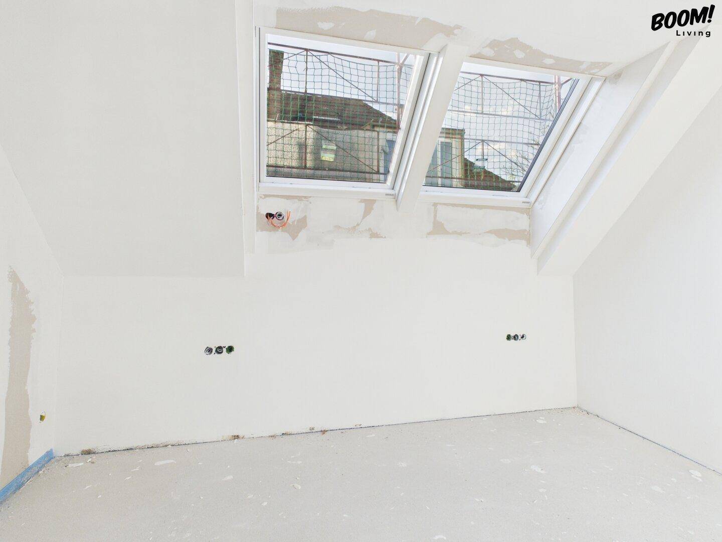 Unfertiger Raum im Dachgeschoss mit Dachfenstern und Blick auf die Dächer.