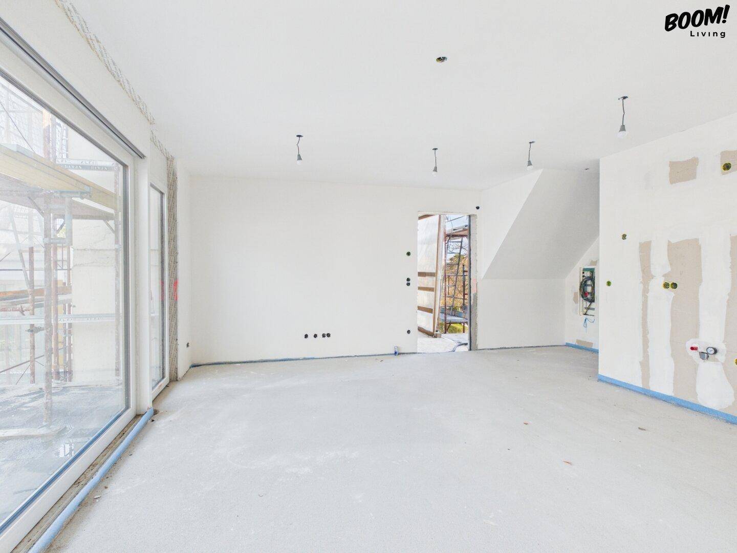 Großer, unfertiger Raum im Rohbau mit breiter Fensterfront und Zugang nach draußen.
