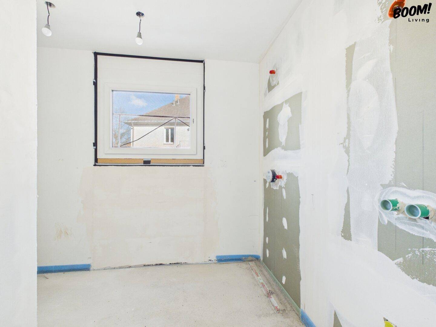 Unfertiger Raum mit Fenster und vorbereiteten Anschlüssen für Sanitär- oder Kücheninstallationen.
