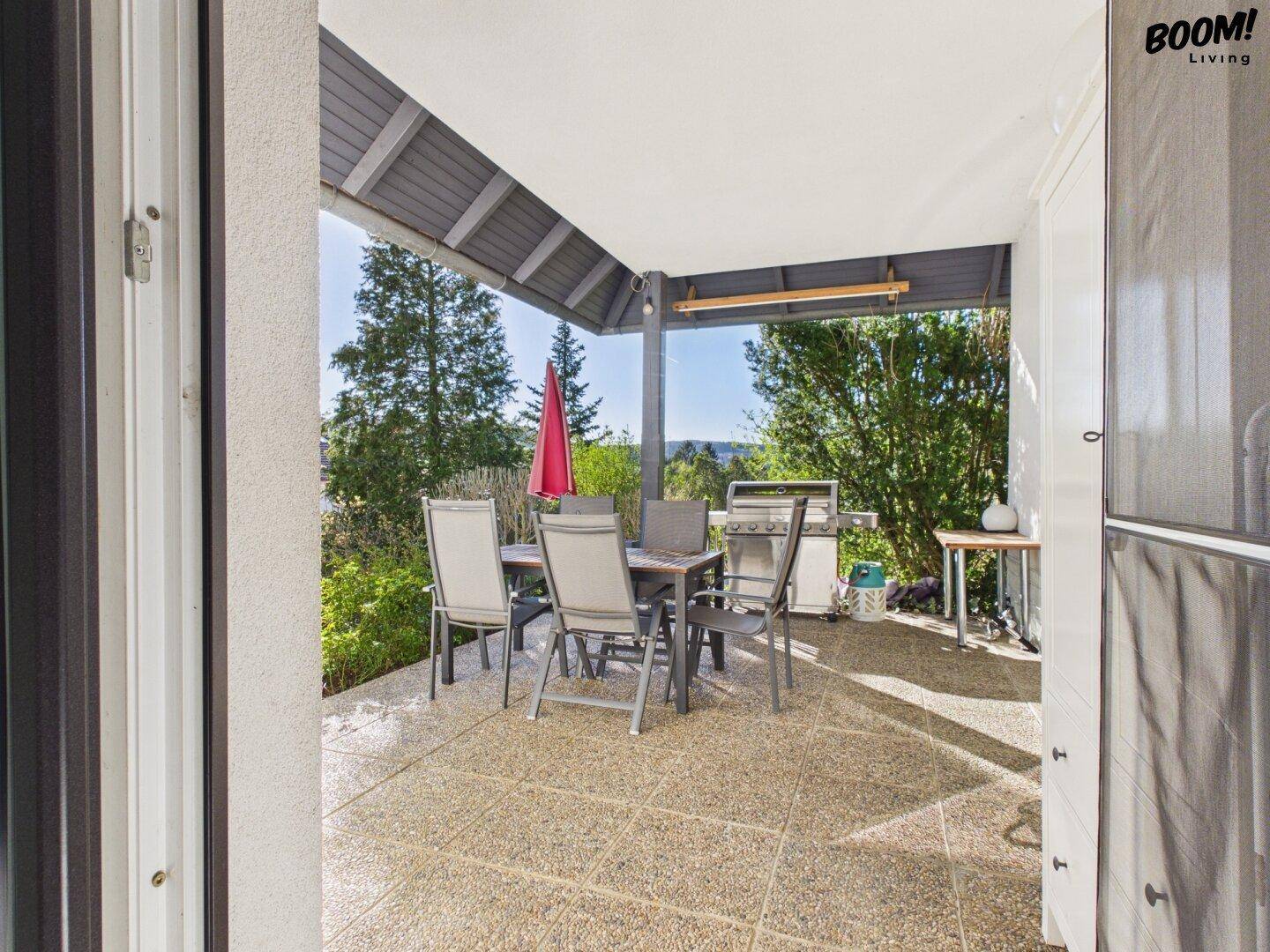 Überdachte Terrasse mit Esstisch, Stühlen und Grill, umgeben von Bäumen.