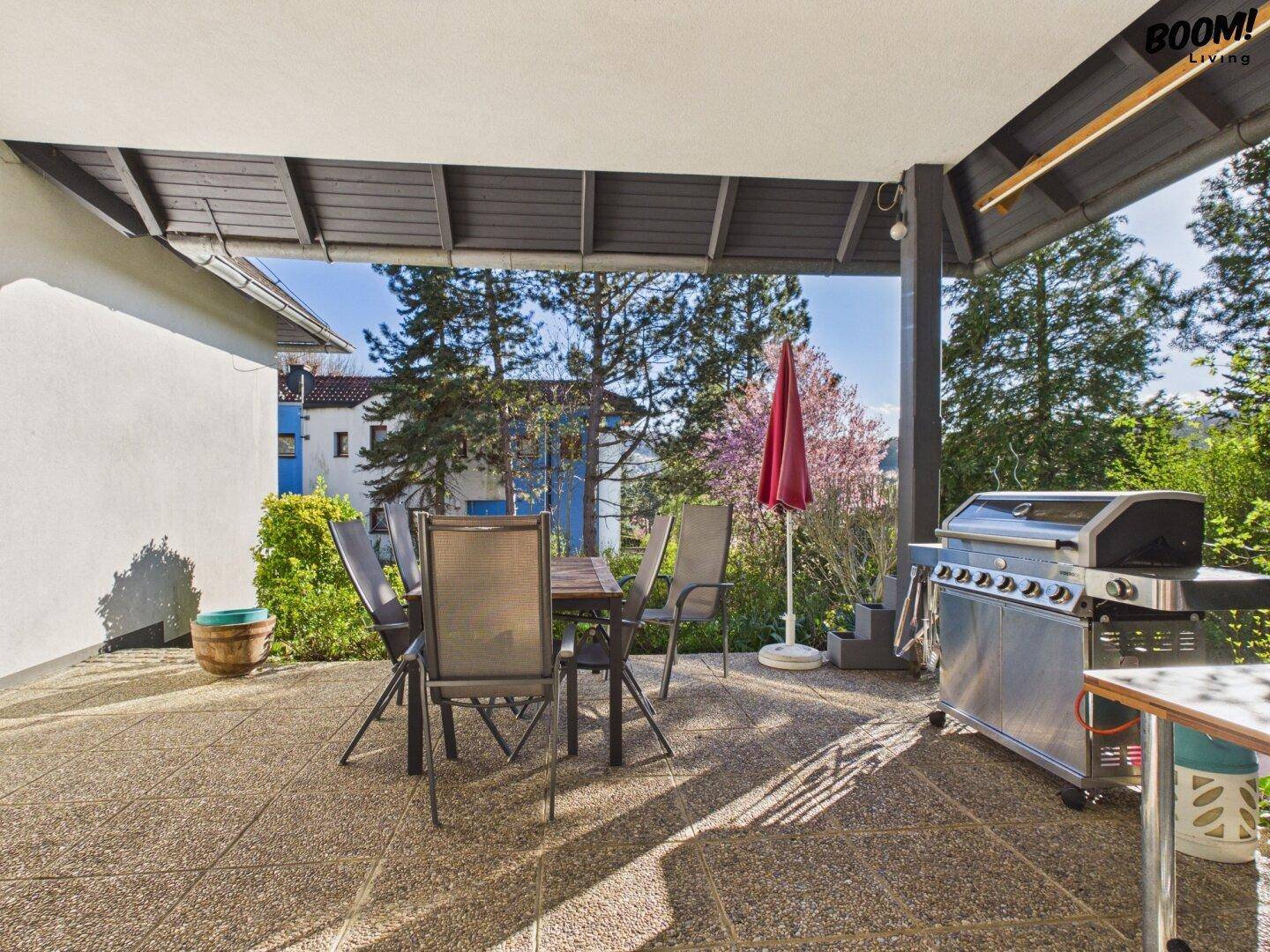 Überdachte Terrasse mit Esstisch, Stühlen und großem Grill, umgeben von Bäumen.