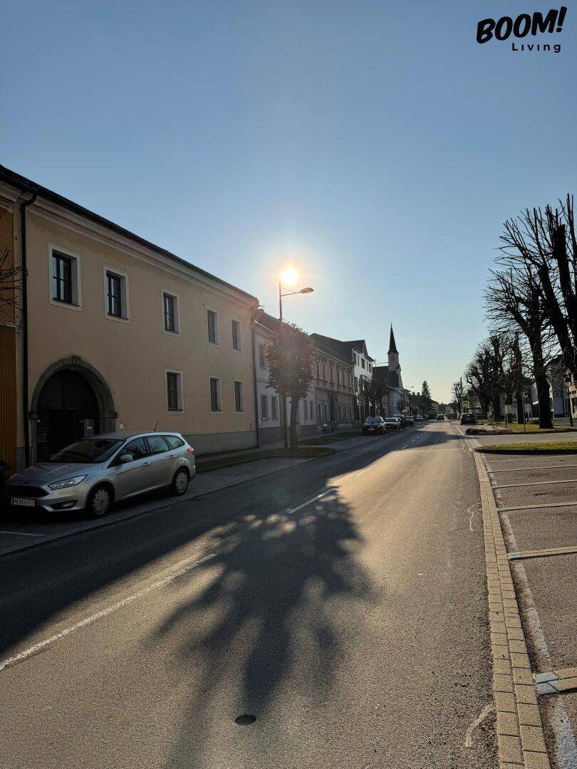 Sonnige Straße mit Gebäuden, Bäumen und einem Kirchturm in der Ferne.