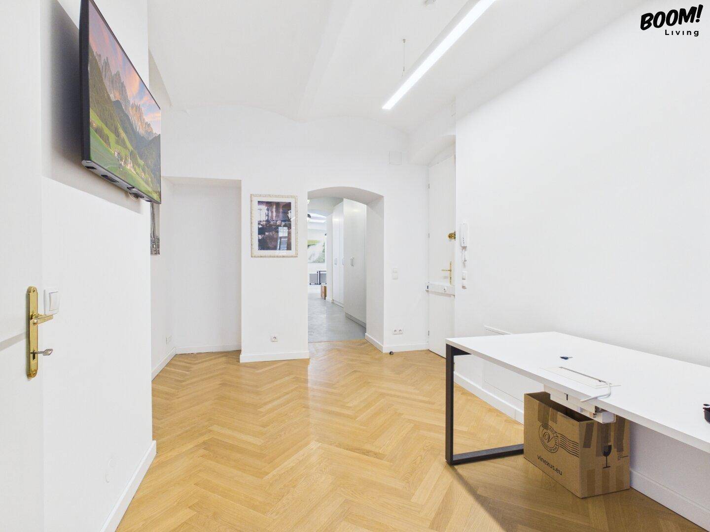 Flur mit Fischgrätparkett, weiß gestrichenen Wänden und einem an der Wand montierten Fernseher.