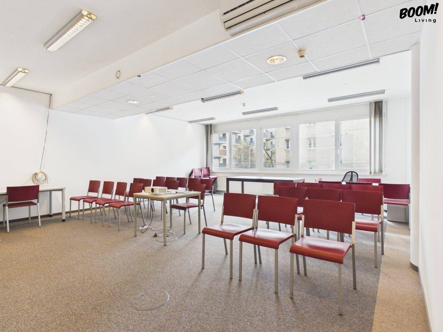 Großer Konferenzraum mit vielen roten Stühlen, Projektor und großen Fenstern für Tageslicht.