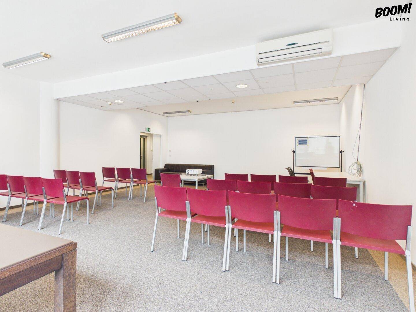 Großer Konferenzraum mit roten Stühlen, Whiteboard und Projektor, ideal für Schulungen und Meetings.