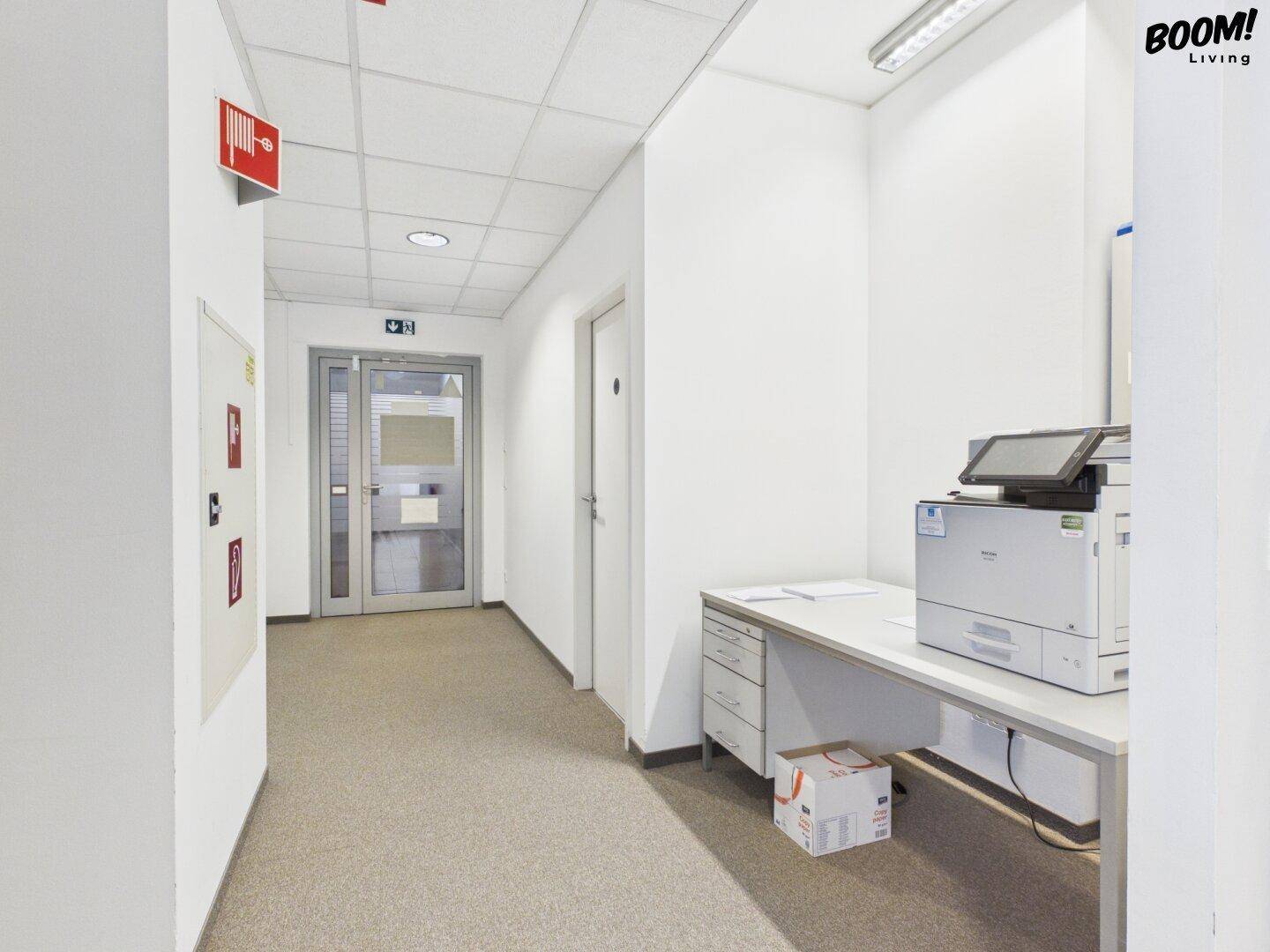 Heller Flur mit Teppichboden, Druckerstation und Brandschutzhinweisen, führt zu weiteren Räumen.