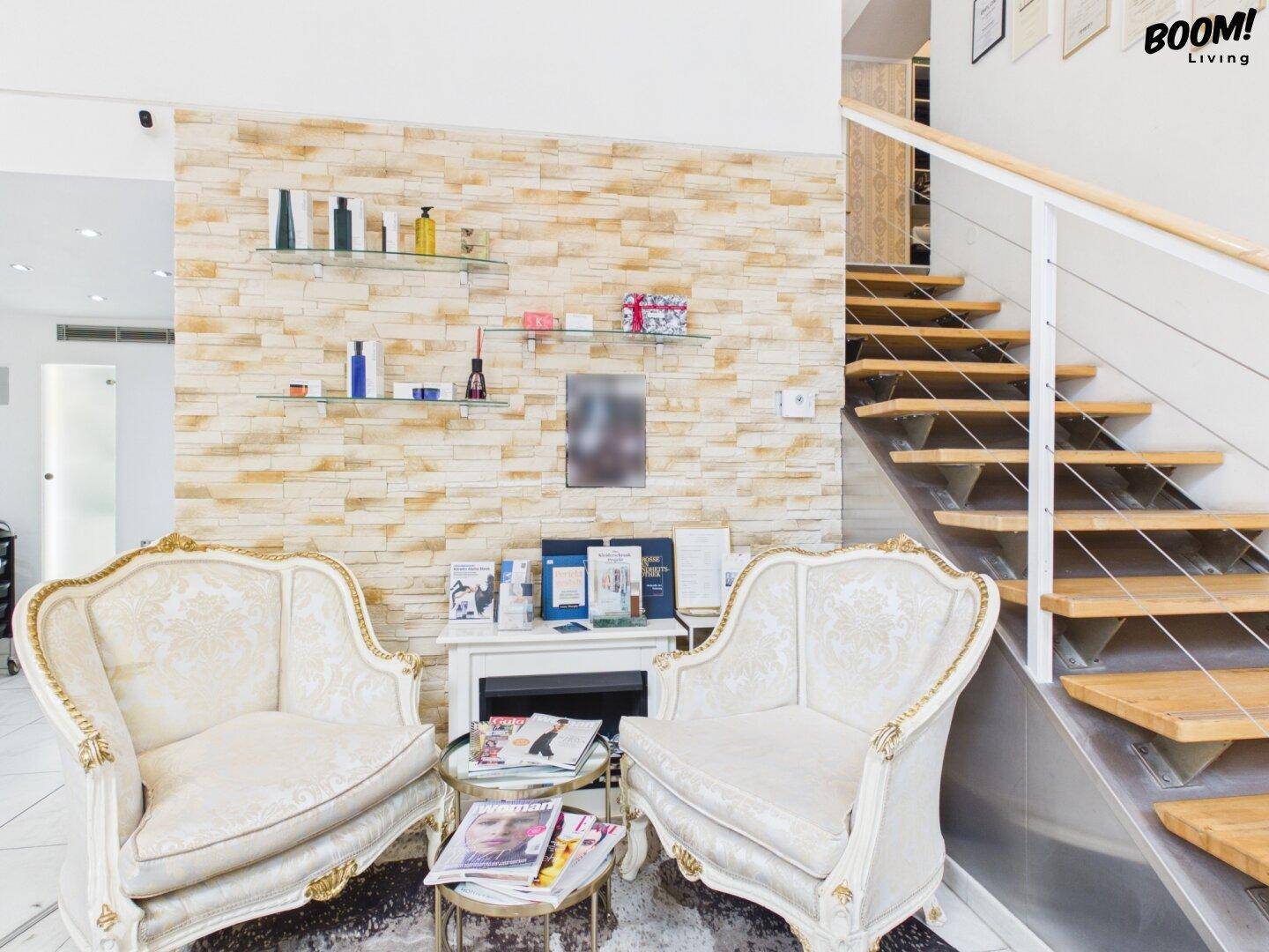 Empfangsbereich eines Schönheitssalons mit zwei eleganten Sesseln, einer Steinwand und einer Holztreppe.