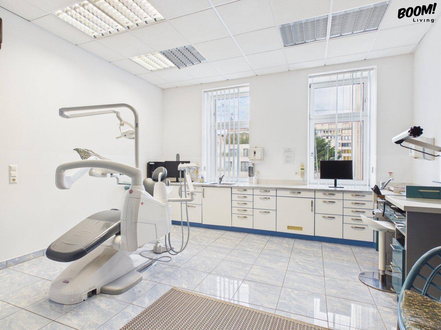 Behandlungszimmer mit Zahnarztstuhl, weißen Schränken und großen Fenstern, hell und funktional eingerichtet.