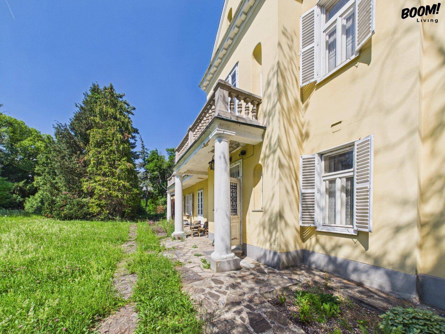 Seitlicher Blick auf die historische Villa mit Terrasse, Säulen und großem Gartenbereich.