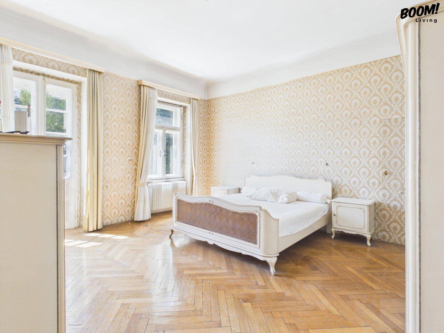 Geräumiges Schlafzimmer mit Fischgrätparkett, gemusterter Tapete und klassischem Bettgestell.