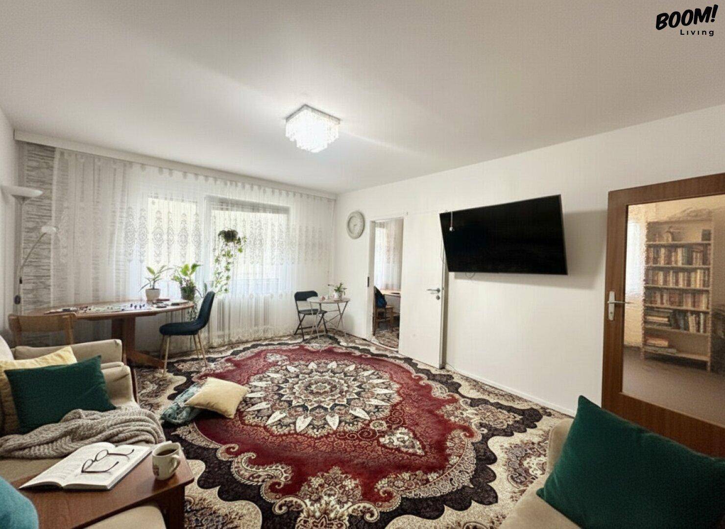 Helles Wohnzimmer mit großem Teppich, Fensterfront und Zugang zu weiteren Räumen der Wohnung.
