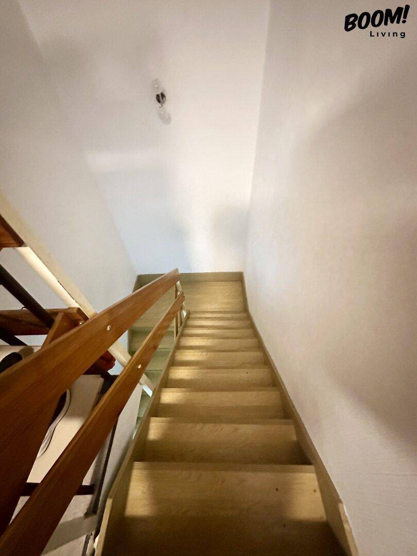Holztreppe führt nach oben, mit schlichten weißen Wänden und einfacher Beleuchtung.