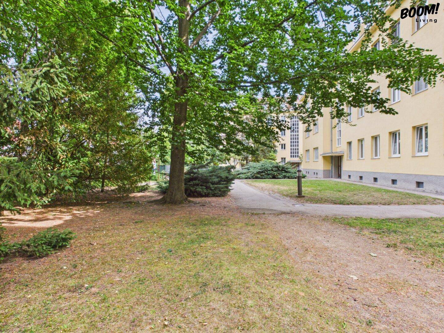 Grüne Außenanlage mit Bäumen, Rasenflächen und einem Weg vor einem Wohngebäude.