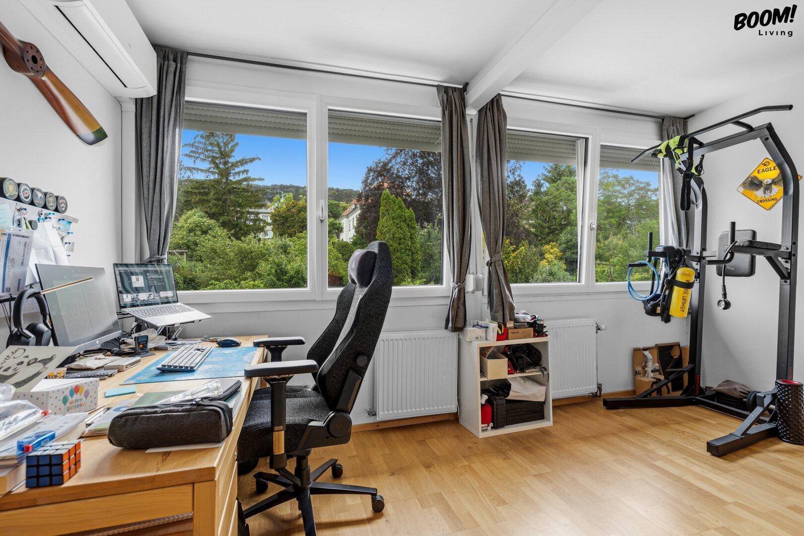 Arbeitszimmer mit Schreibtisch, Sportgeräten und großen Fenstern mit Blick ins Grüne.