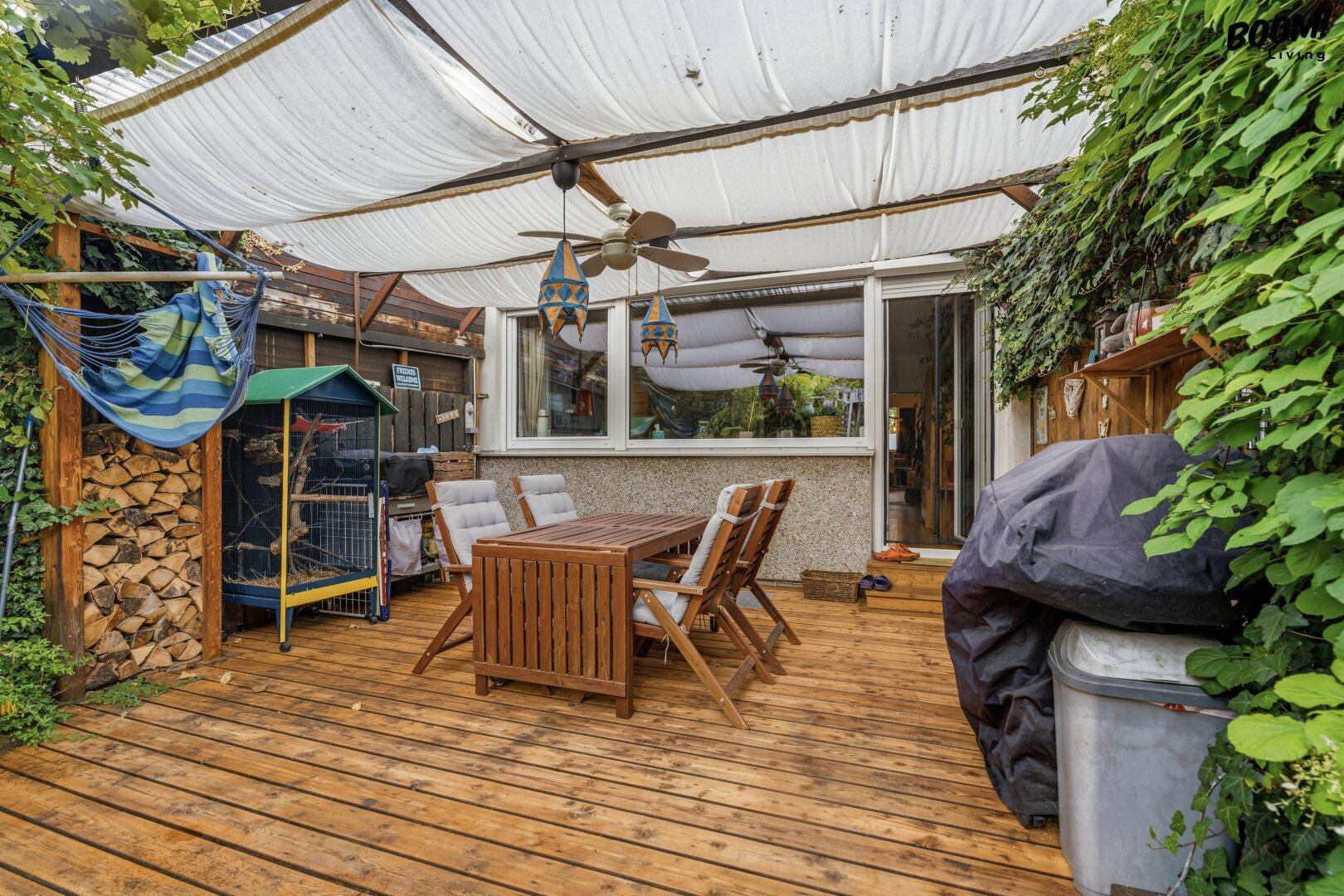 Geräumige Holzterrasse mit Essbereich, Holzstapel und überdachtem Grill, umgeben von Grün.