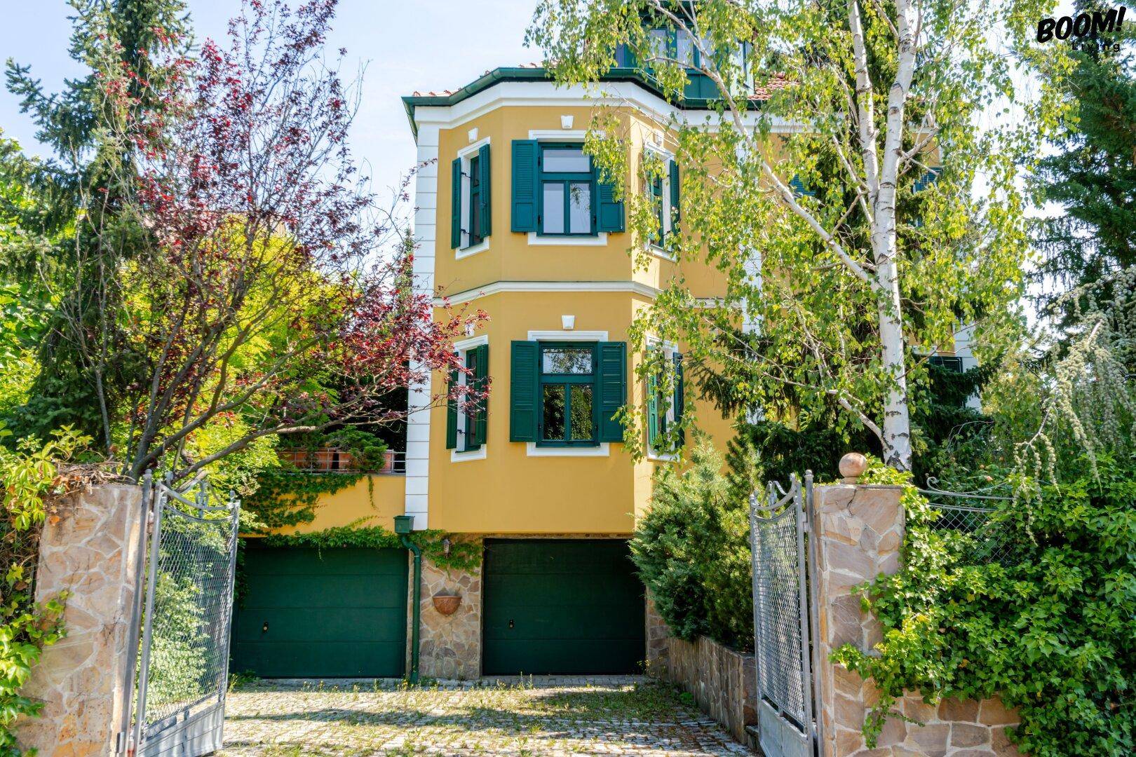 Vorderansicht der Villa mit gelber Fassade, grünen Garagentoren und umzäuntem Eingangsbereich.