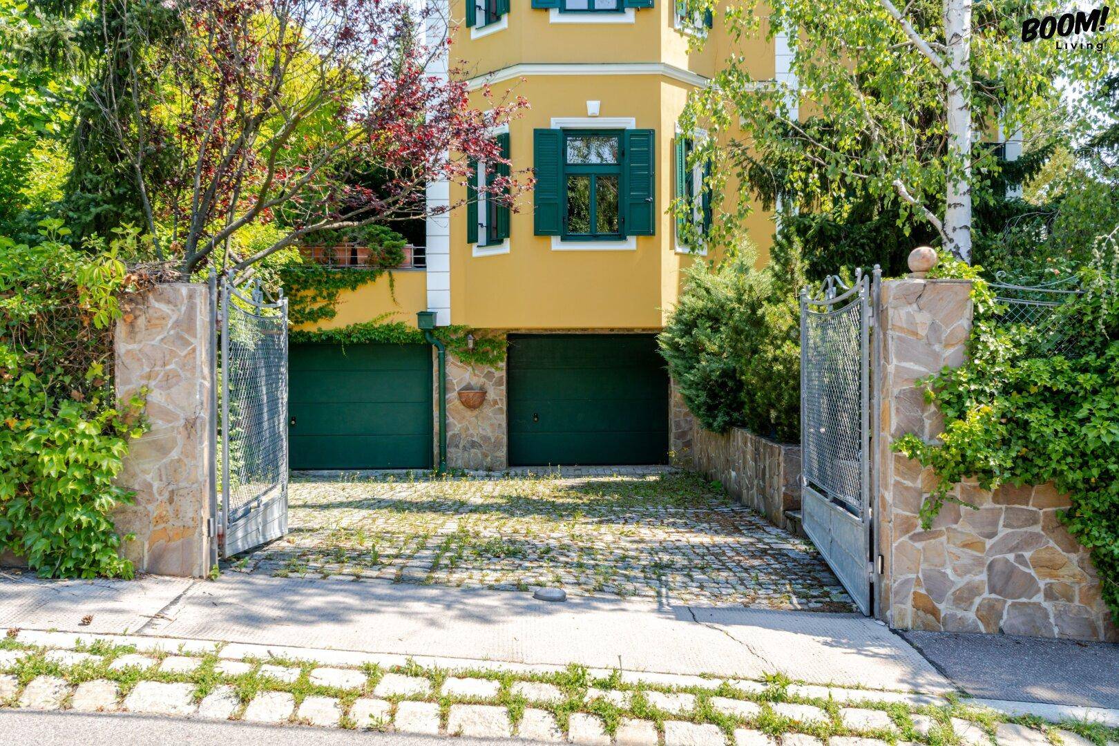 Gelbes Gebäude mit grünen Fensterläden und Garagentoren, umgeben von üppigem Grün und einem gepflasterten Hof.