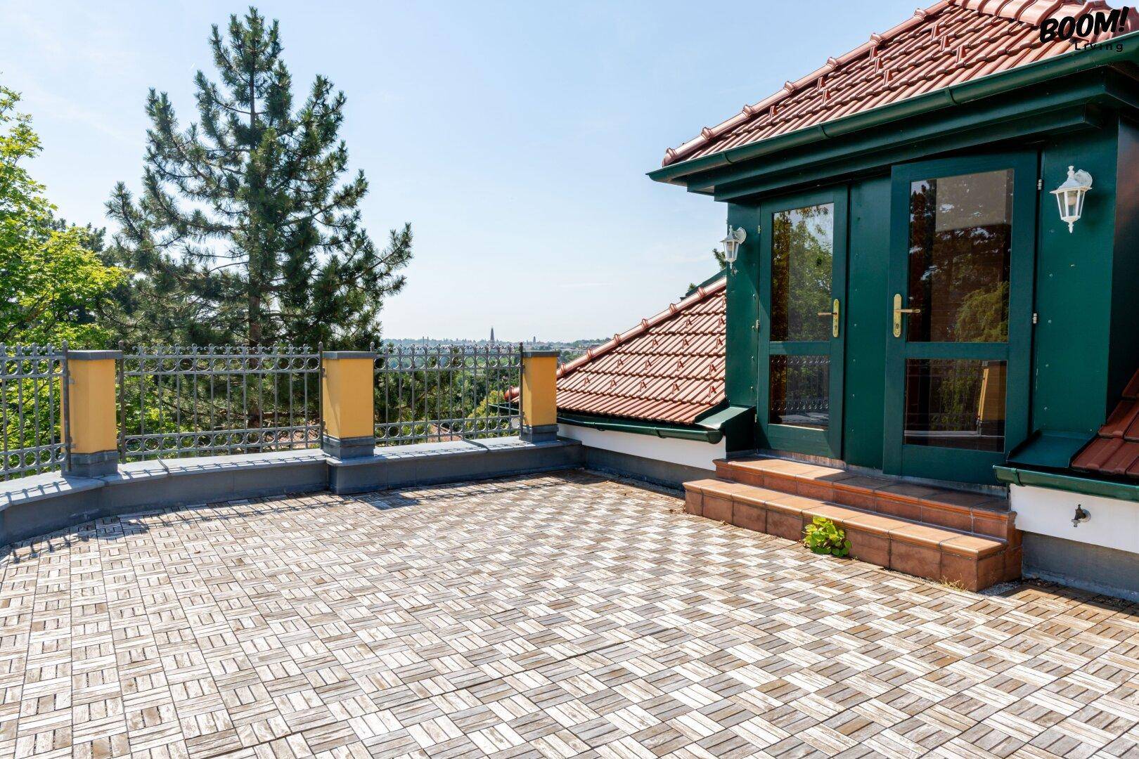 Helle Terrasse mit Holzboden und Zugangstür, umgeben von einem Metallgeländer und Bäumen.