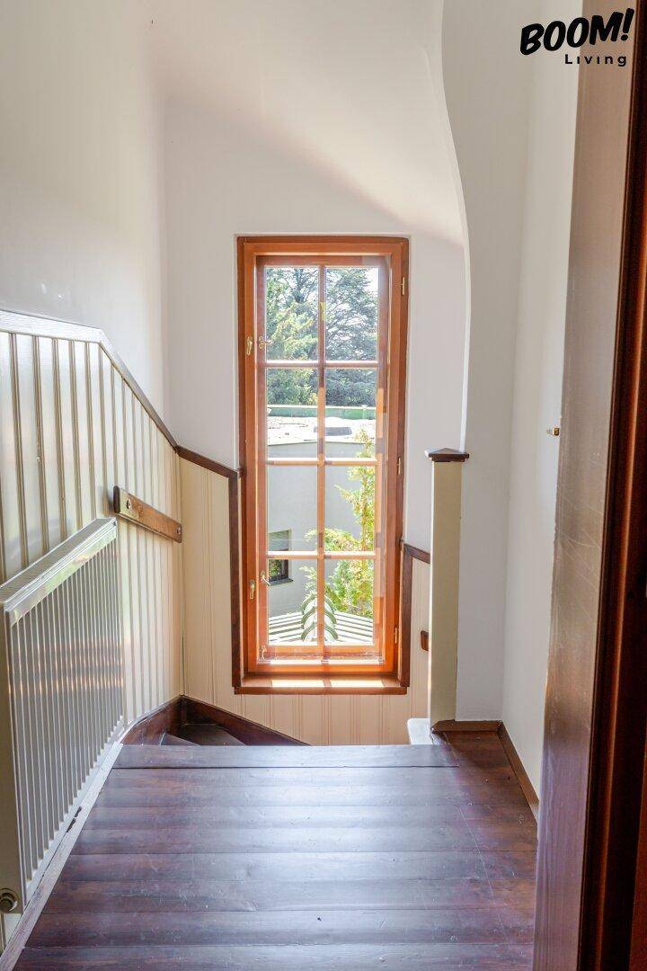 Holztreppe mit hellem Geländer und einem hohen Fenster, das viel Tageslicht hereinlässt.