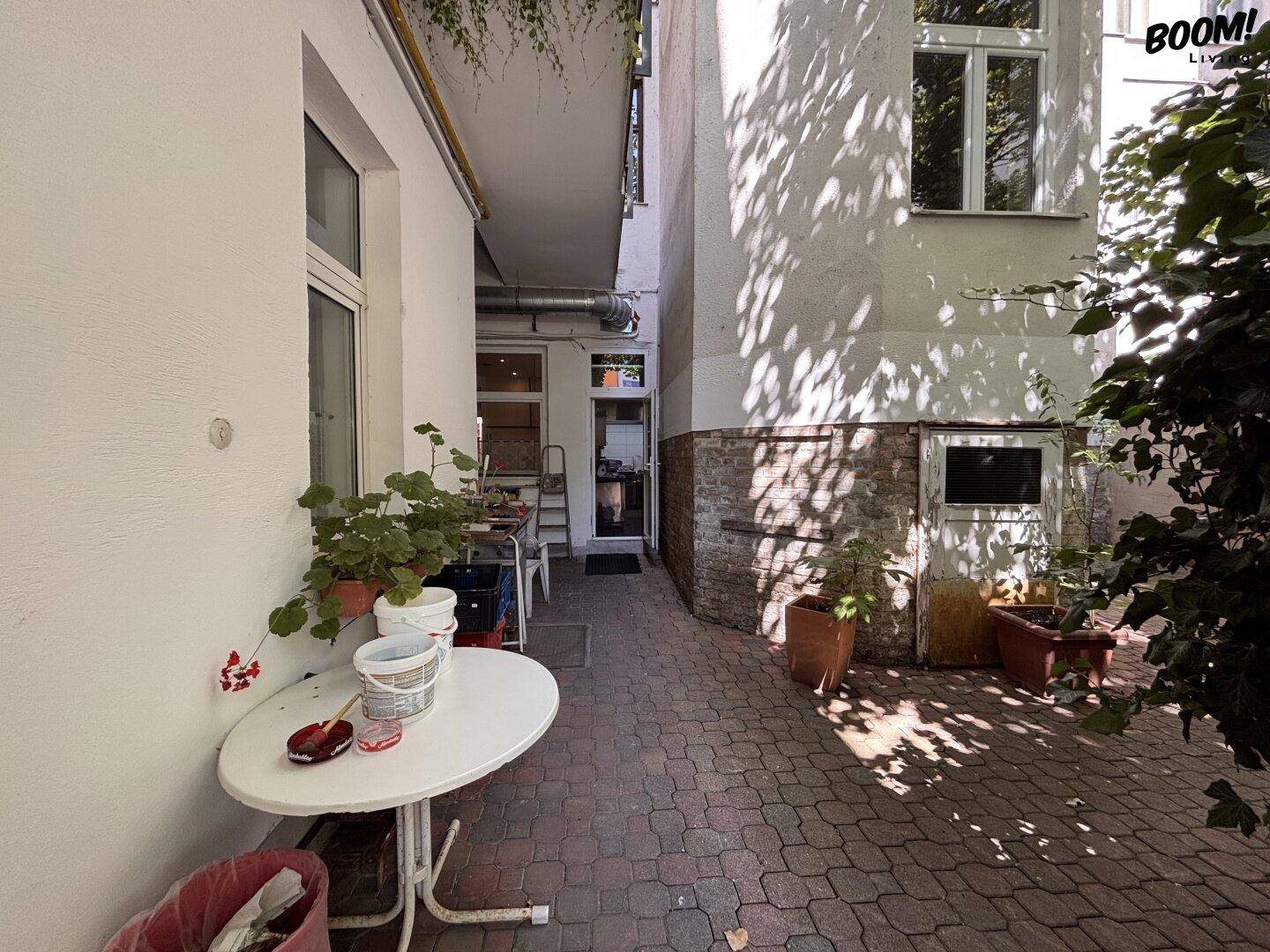 Gepflasterter Außenbereich zwischen Gebäuden mit Tischen, Pflanzen und einem schmalen Durchgang.