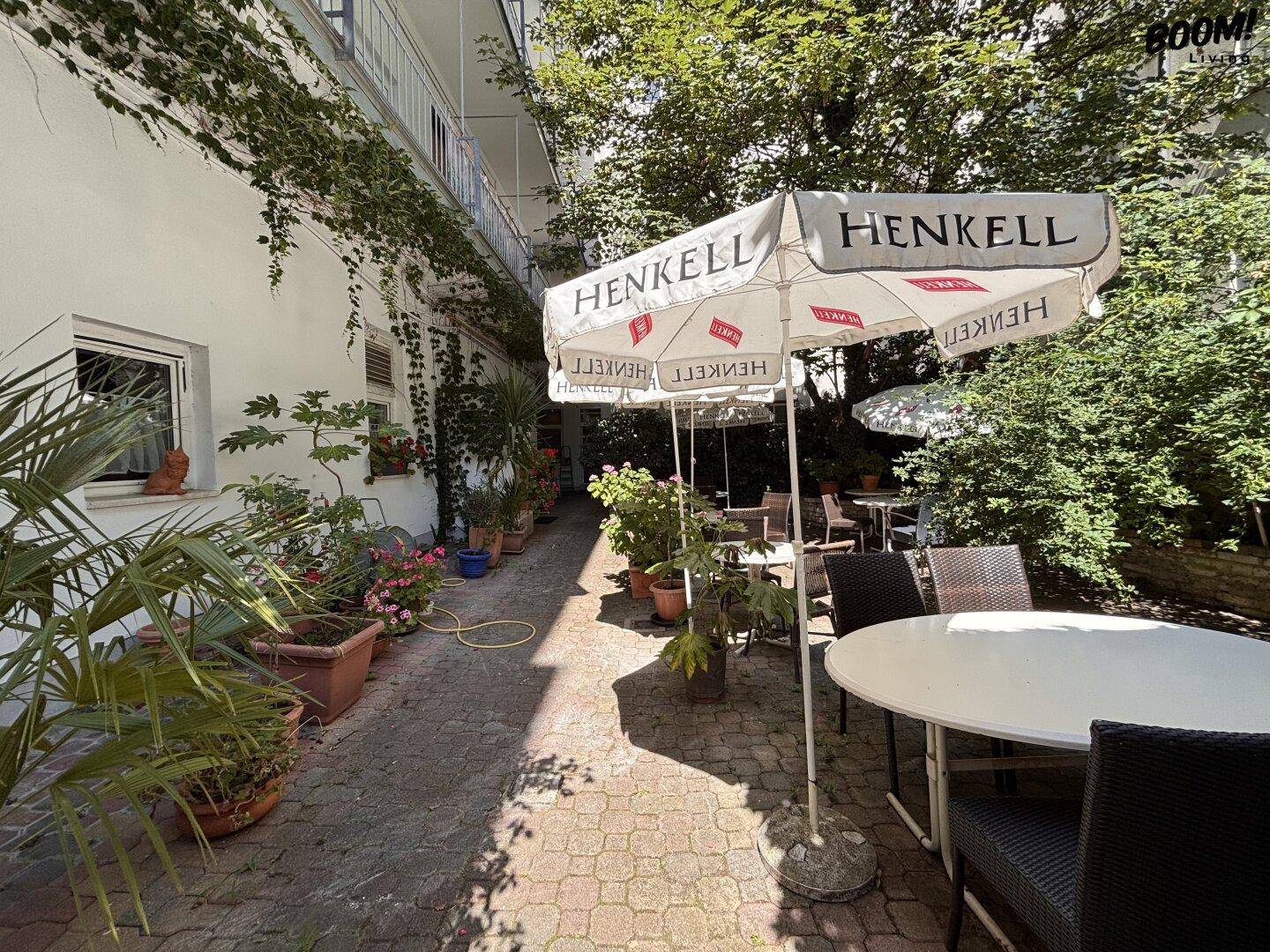 Lange, gepflasterte Terrasse mit vielen Pflanzen, Tischen und Sonnenschirmen entlang einer Gebäudewand.