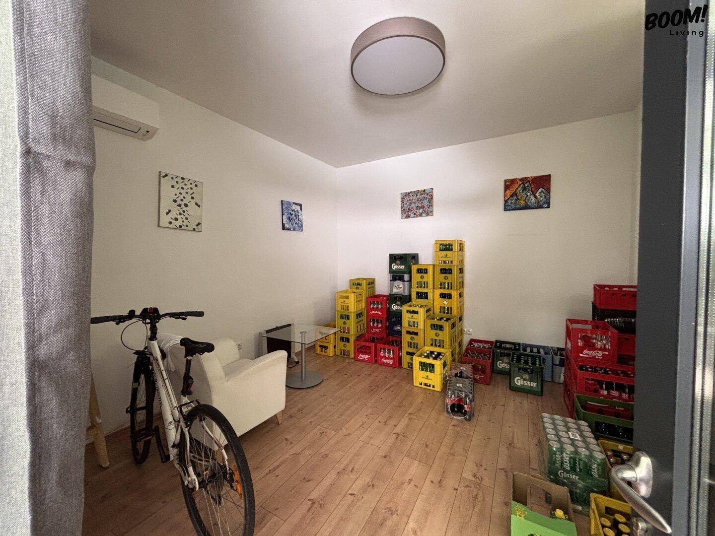 Lagerraum mit Holzboden, Klimaanlage und gestapelten Getränkekisten, auch ein Fahrrad ist sichtbar.