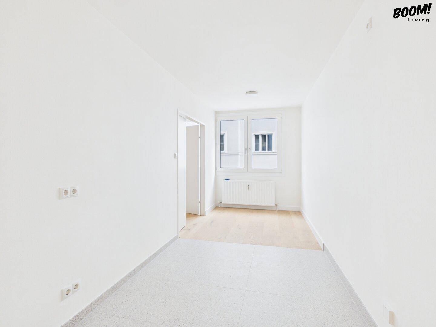 Leerer Raum mit hellen Wänden, einem Fenster und einem Übergang von Fliesen zu Parkettboden.