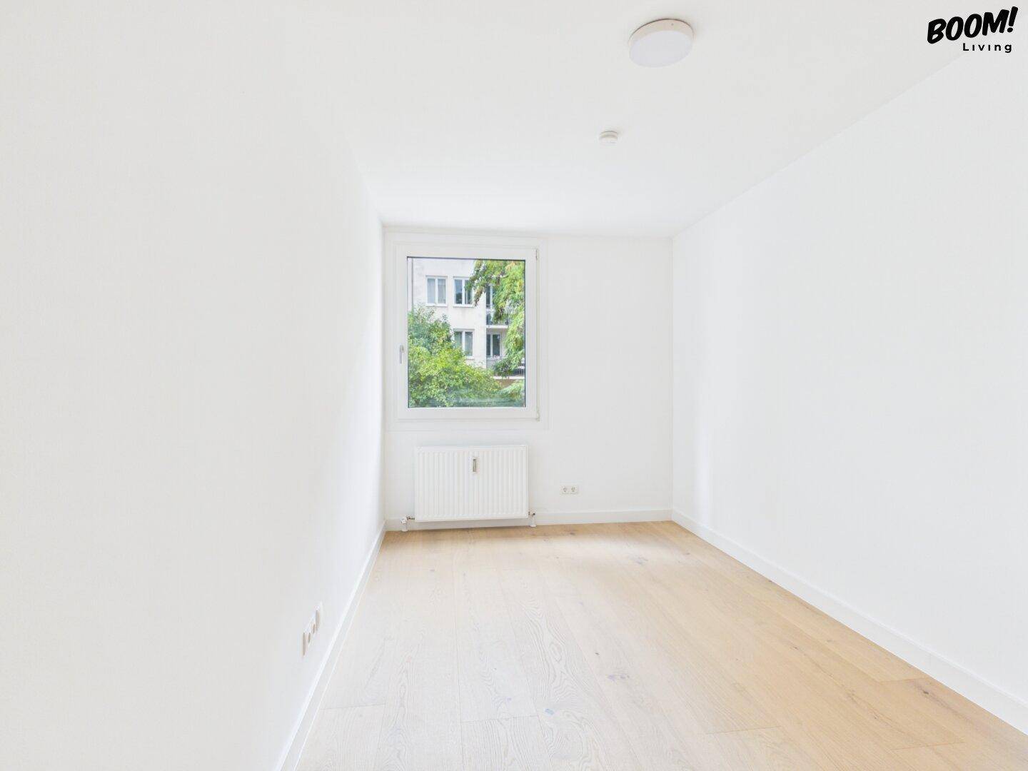 Helles Schlafzimmer mit Parkettboden, weißen Wänden und einem Fenster mit Blick ins Grüne.