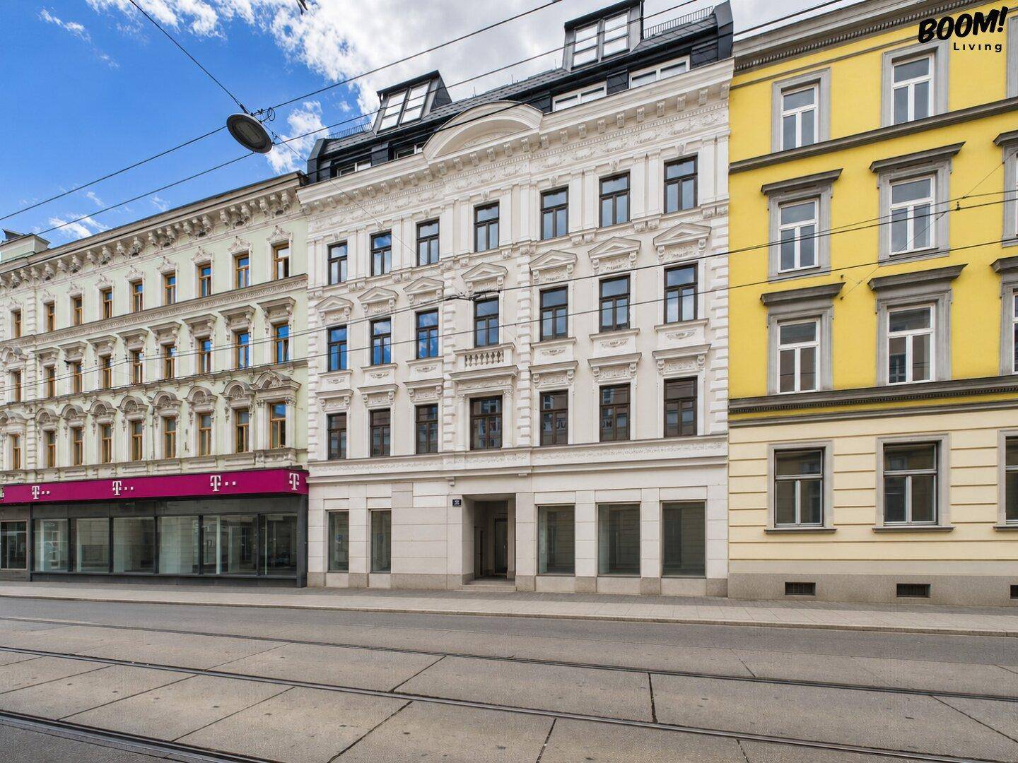 Straßenansicht eines historischen Gebäudes mit reich verzierter weißer Fassade und Dachausbau, umgeben von weiteren Gebäuden.