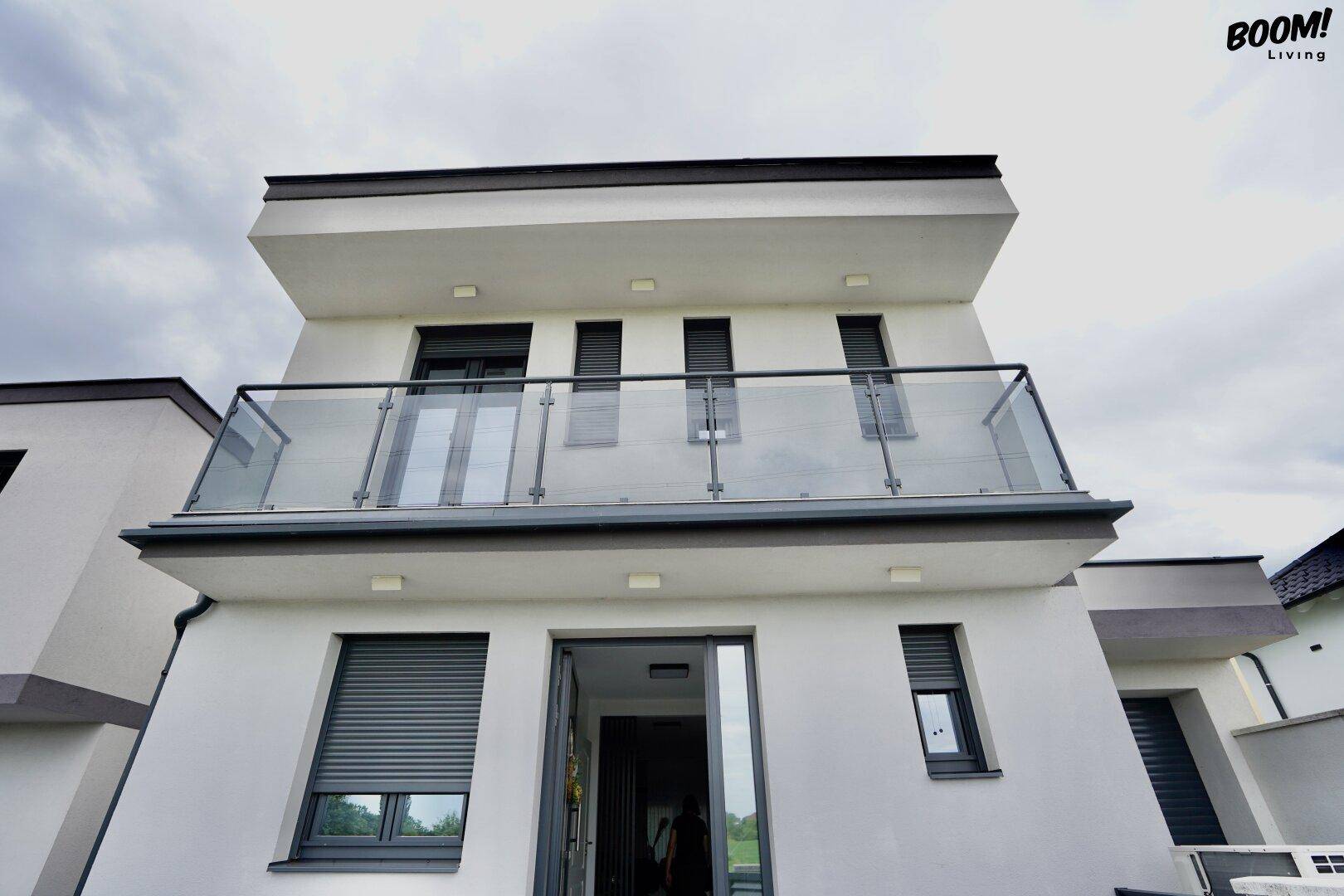 Moderne Hausfassade mit Balkon und Glasgeländer sowie Fenstern mit Rollläden.