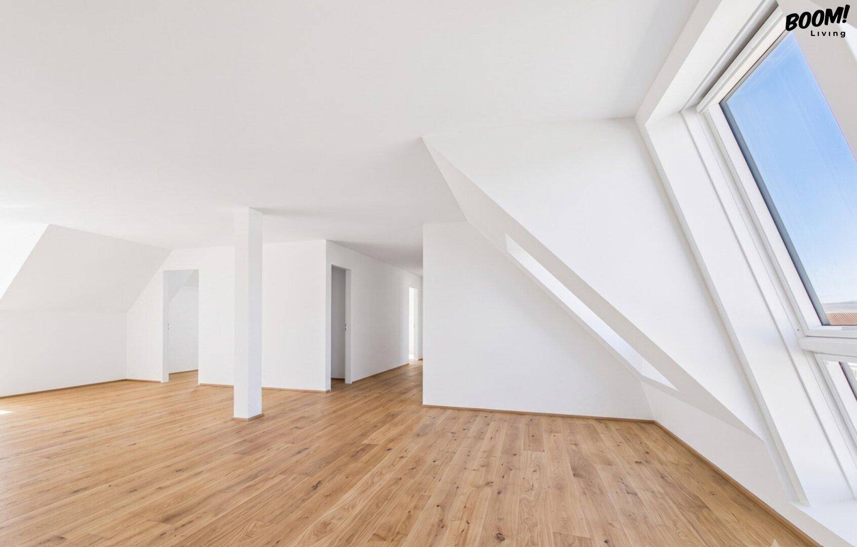 Geräumiger, heller Dachgeschossraum mit Holzboden, weißen Wänden und schrägen Fenstern.