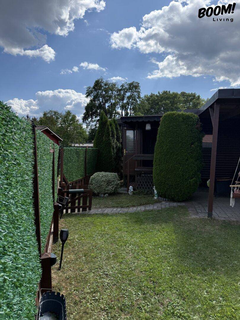 Sonniger Garten mit Rasenfläche, hohen Hecken und einem charmanten Holzhaus mit überdachtem Bereich.