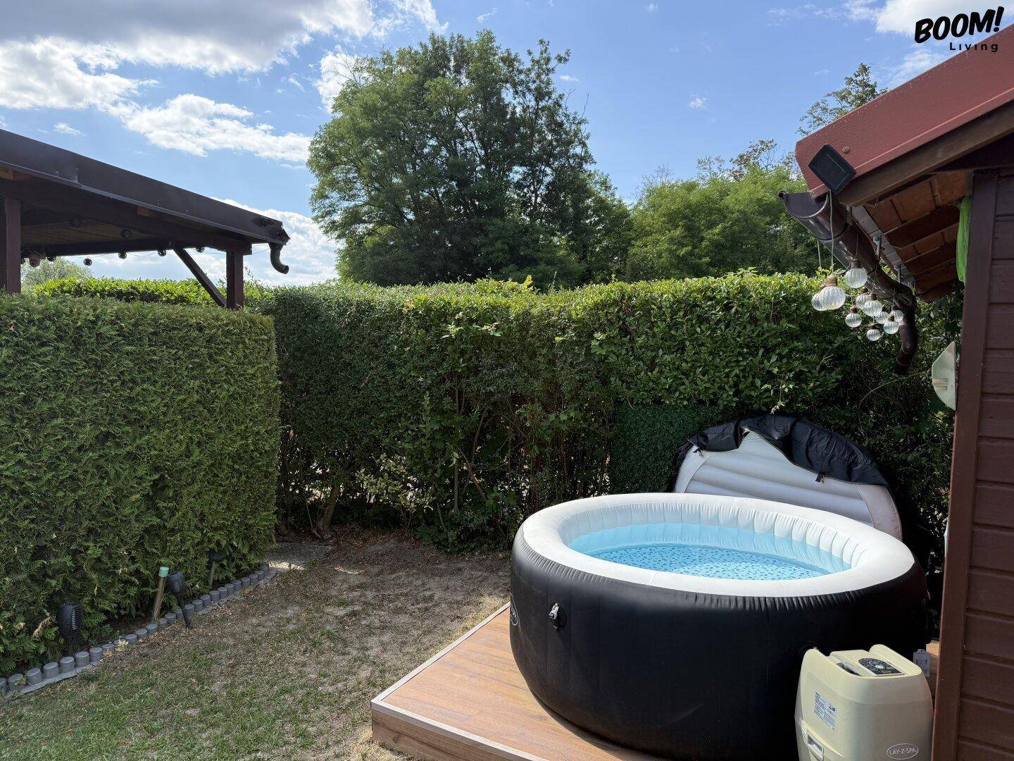 Aufblasbarer Whirlpool auf einer Holzterrasse, umgeben von einer dichten grünen Hecke unter blauem Himmel.