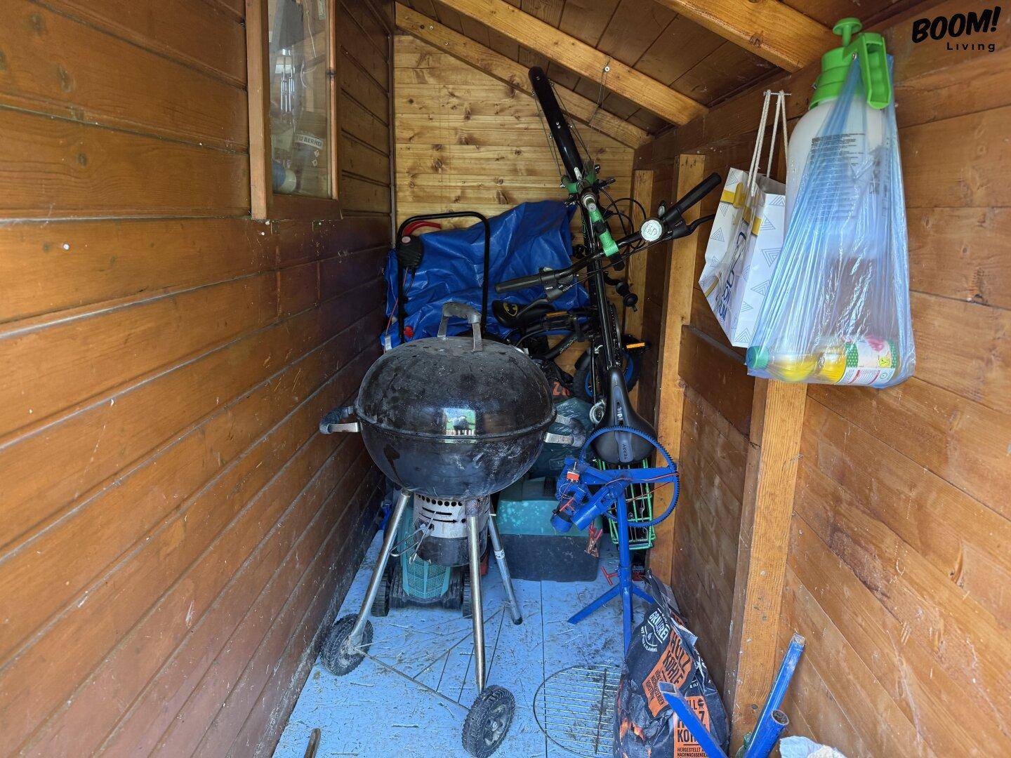 Innenansicht eines Holzschuppens mit einem Grill und verschiedenen Gartengeräten und Lagergegenständen.