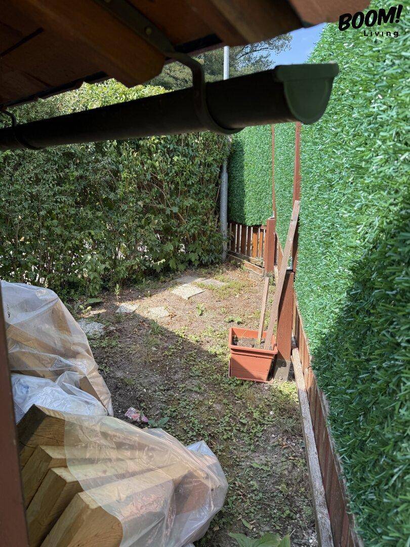 Schmaler Gartenweg zwischen einer natürlichen Hecke und einem grünen Sichtschutzzaun mit gelagertem Holz.