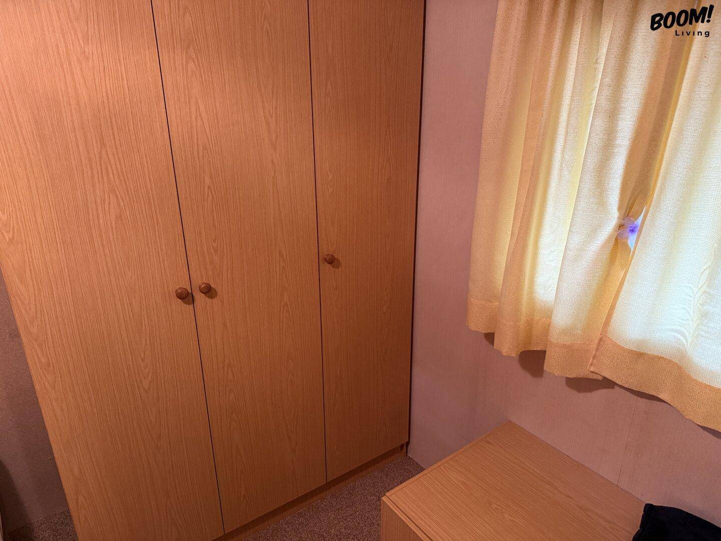 Kleiderschrank aus hellem Holz und ein Fenster mit gelben Vorhängen in einem einfach eingerichteten Raum.