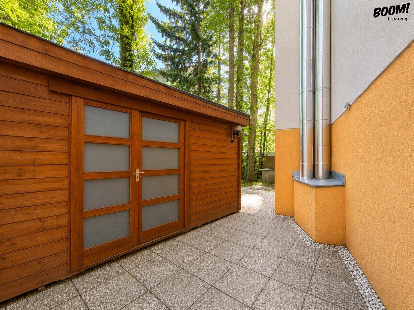 Hölzernes Gartenhaus mit großen Fenstertüren und gepflastertem Vorplatz im Grünen.