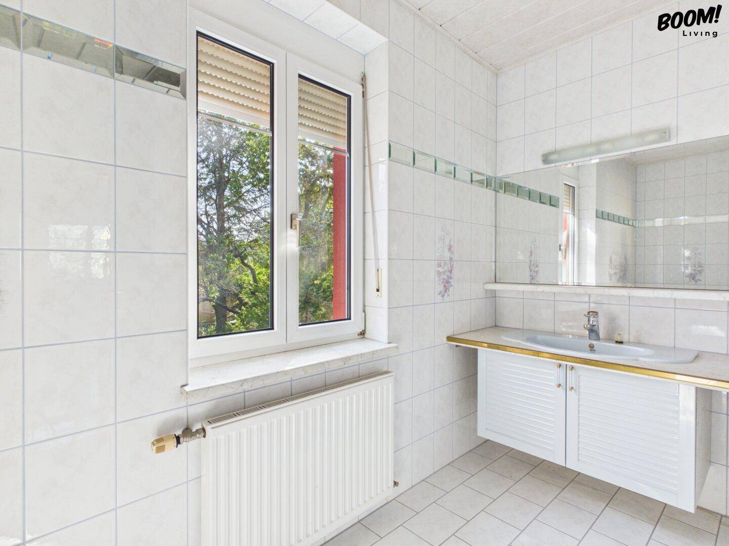 Badezimmer mit Fenster, Waschbecken und Spiegel, komplett gefliest in hellem Weiß.