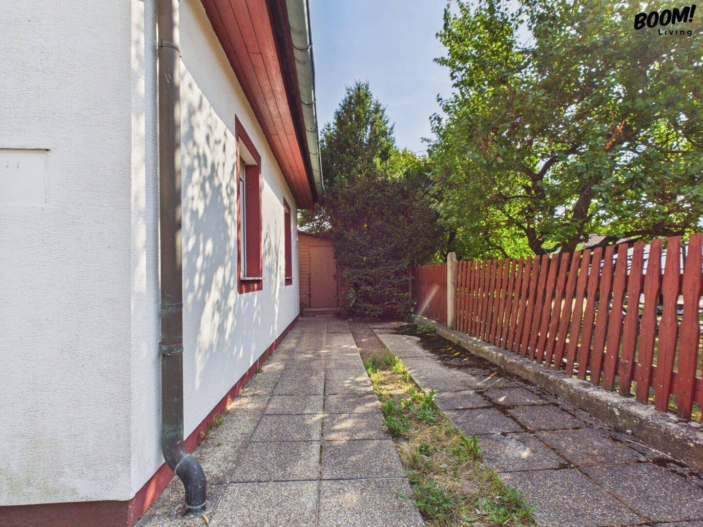 Seitenansicht des Hauses mit gepflastertem Weg und Holzzaun, umgeben von Bäumen.