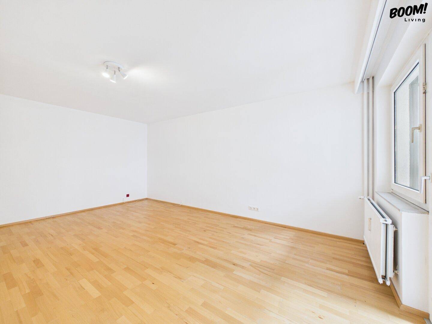 Leeres Zimmer mit Parkettboden und Fenster, das viel Tageslicht hereinlässt, ideal als Schlafzimmer.