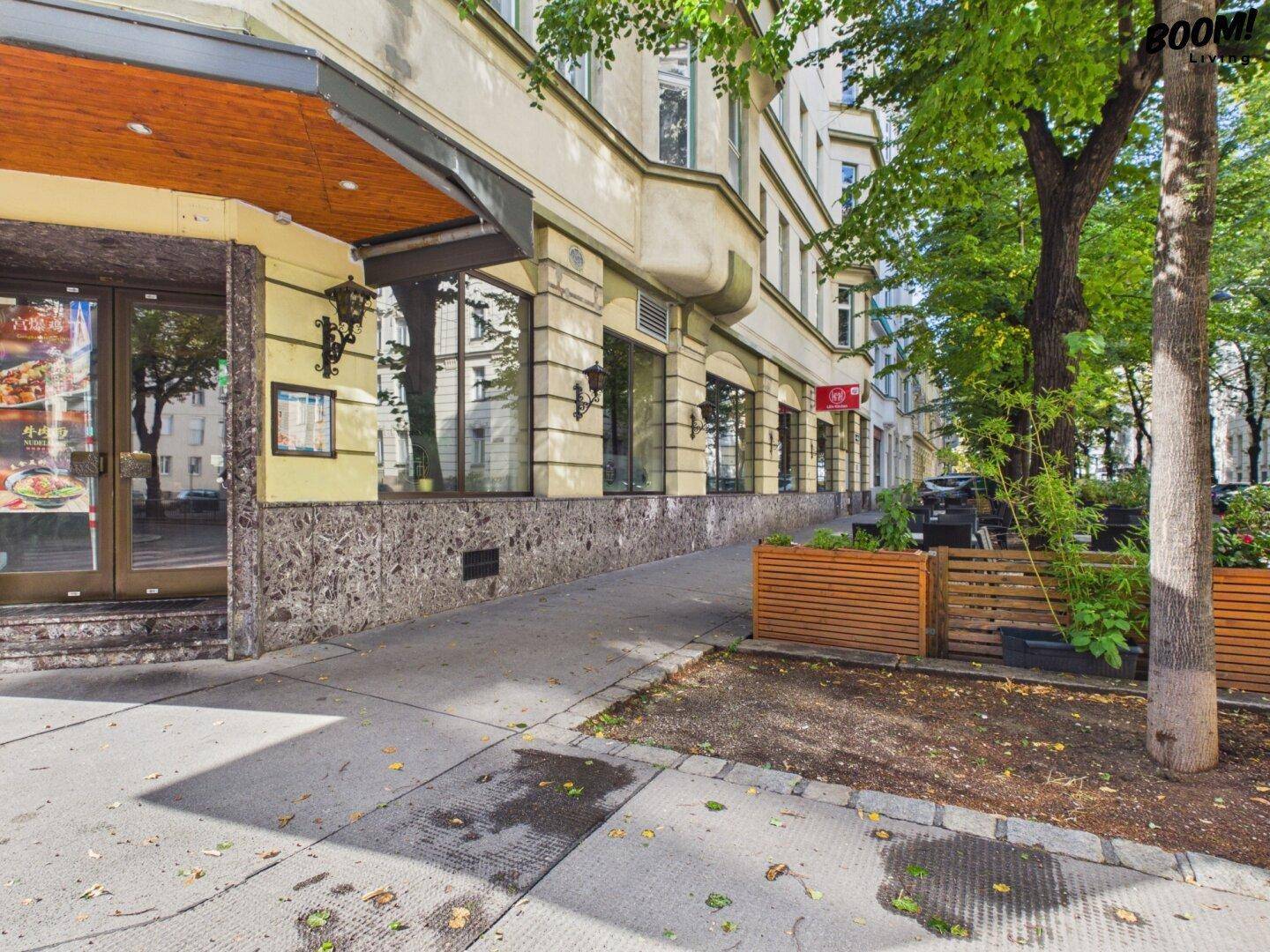 Straßenszene mit Geschäftsfassaden im Erdgeschoss und Bäumen am Gehweg.