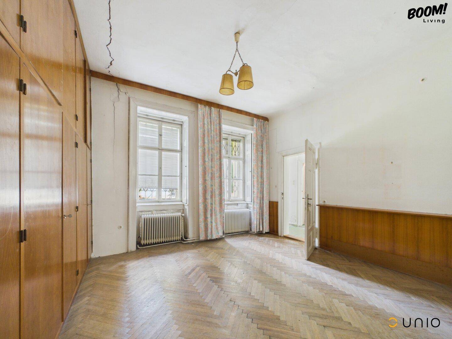 Schlafzimmer mit Fischgrätparkett, Einbauschränken und zwei Fenstern mit Vorhängen.