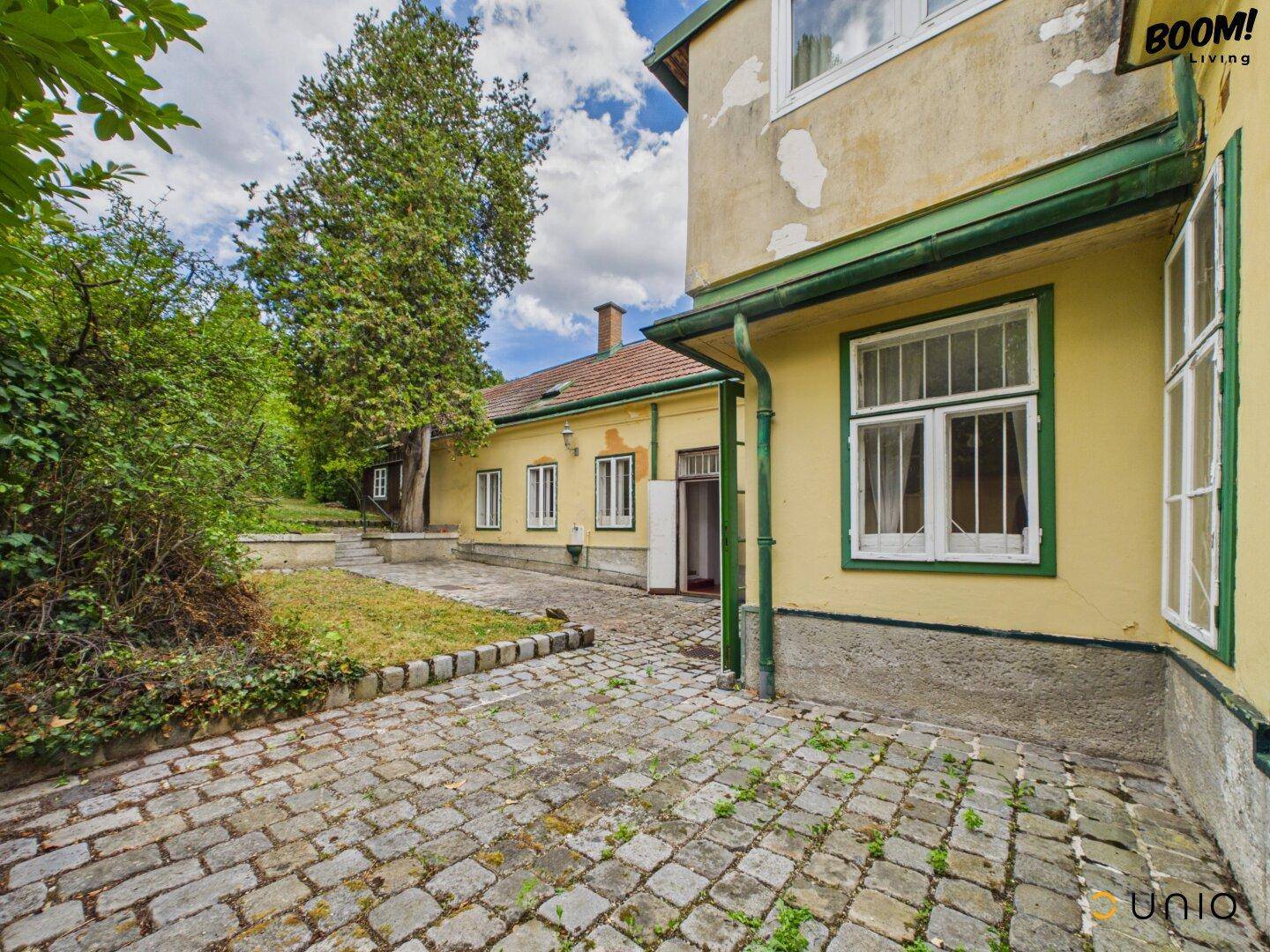 Historisches Gebäude mit gelber Fassade, grünen Details und gepflastertem Außenbereich.