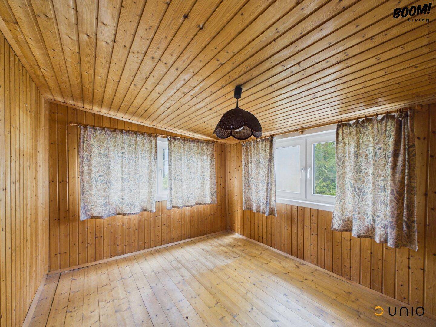 Gemütlicher Raum mit Holzverkleidung, Holzdielenboden und Fenstern mit gemusterten Vorhängen.