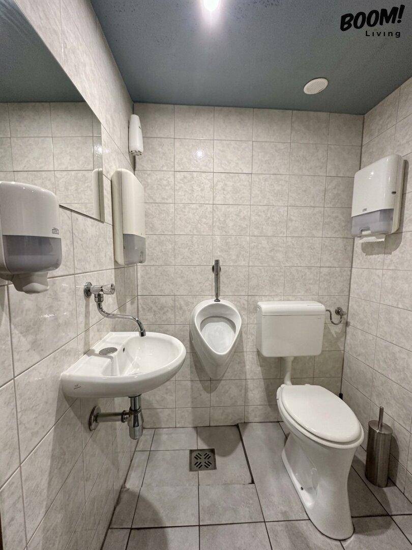 Öffentliche Toilette mit Urinal, Waschbecken und gefliesten Wänden in hellem Farbton.