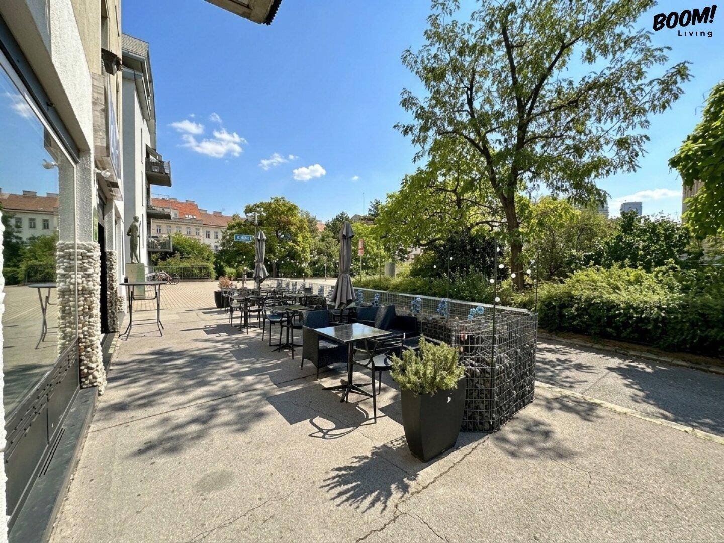 Sonniger Außenbereich mit Sitzgelegenheiten und Sonnenschirmen, umgeben von viel Grün und Bäumen.