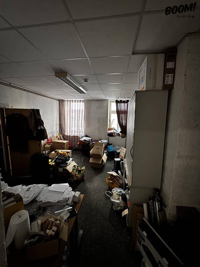 Unaufgeräumtes Büro oder Lagerraum mit Teppichboden, abgehängter Decke und zwei Fenstern mit Vorhängen.