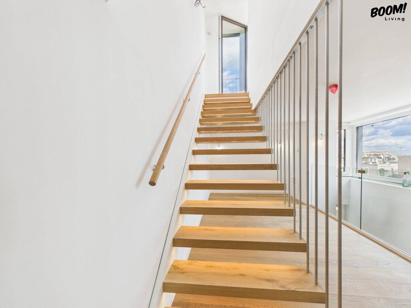 Moderne Holztreppe mit Metallgeländer, die zu einem oberen Bereich mit viel Tageslicht führt.