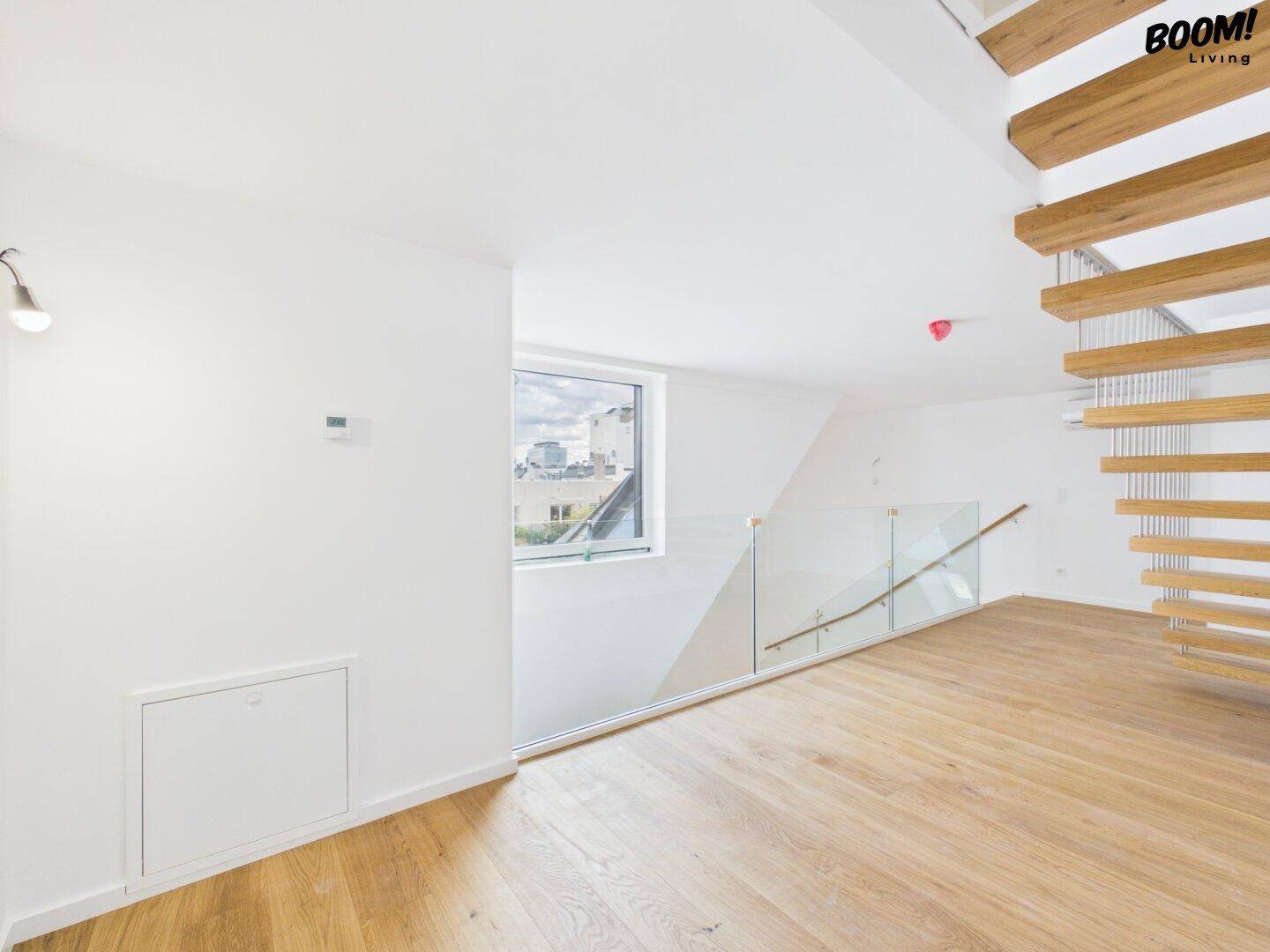 Heller Raum mit Holzfußboden, großem Fenster und Stadtblick, verbunden mit einem Glasgeländer.