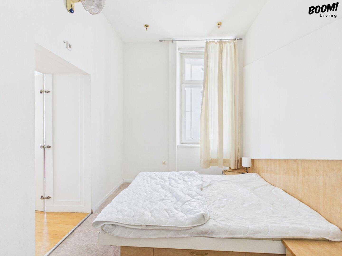 Schlichtes Schlafzimmer mit Doppelbett, Holzkopfteil und Fenster mit Vorhängen.